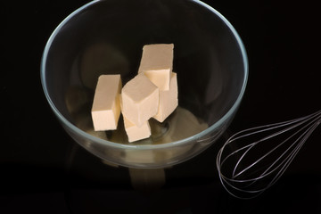 Pieces of butter lie in a glass bowl on a dark background in the studio.  Nearby is a metal whisk...