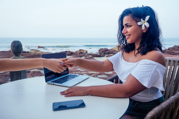 young Indian Asian woman in a business meeting (interview) with a caucasian white woman in tropical paradise beach. multinational handshake and high five successful deal ocean restaurant background