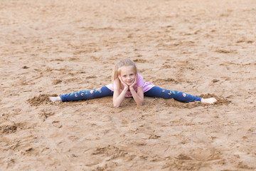 Little blonde girl stretches on sand outdoors