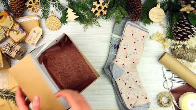 Female woman adult in beige warm sweater preparing gift present socks with christmas ornament on wooden background with pine tree branches. Merry Christmas Eve and Happy New Year concept.
