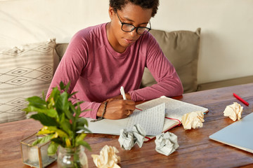 Cropped image of college student with dark skin, piercing does homework at workplace, writes down...