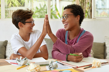 Glad multiethnic dark skinned woman and youngster give high five to each other, sit at workplace,...