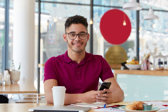 Handsome cheerful young man searches job, browses webpage on modern cell phone, checks information in internet, sits at cafeteria, enjoys sweet dessert and fresh drink, looks gladfully at camera