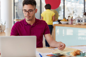 Busy male employer does remote work on laptop computer, downloads files from internet, wants to have successful career, surrounded with papers, wears glasses for vision correction. Technology concept