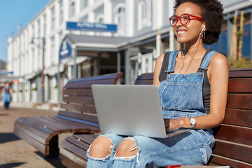 Positive Afro American woman listens music with earphones and works on laptop laptop, dressed in denim overalls, smiles positively, sits at bench against urban environment, focused into distance