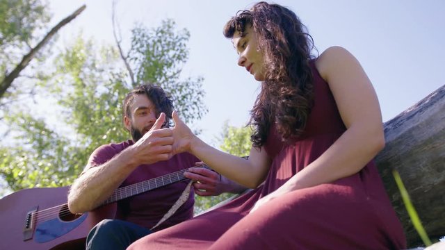 low angle handheld of a young couple playing thumb wars in a park on a fallen tree, male holding his guitar