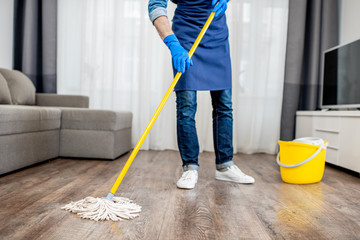 Man as a professional cleaner in blue uniform washing floor with mopping stick and bucket in the...