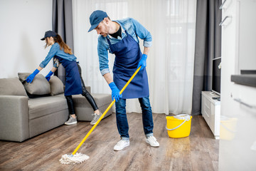 Professional cleaners in blue uniform washing floor and wiping dust from the furniture in the...