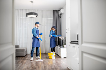 Professional cleaners in blue uniform washing floor and wiping dust from the furniture in the living room of the apartment. Cleaning service concept