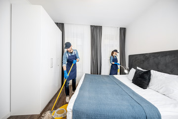 Professional cleaners in uniform washing floor and wiping dust from the furniture in the hotel room...