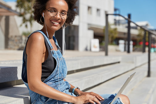 Image of happy delighted Afro American woman keyboards information on laptop computer, connected to wireless 4G internet, sits outside in big city with free space for your promotion or slogan