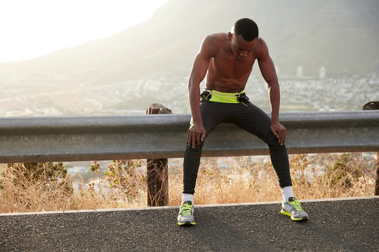 Tired black sportsman breathes with difficulty, rests with arms on knees, being fatigue after racing competition, wears leggings and comfortable shoes, copy space against mountain landscape.