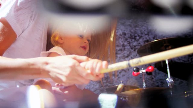 Cute blue eyed child supported by attractive young Caucasian mother while playing with drumsticks and drums home studio 