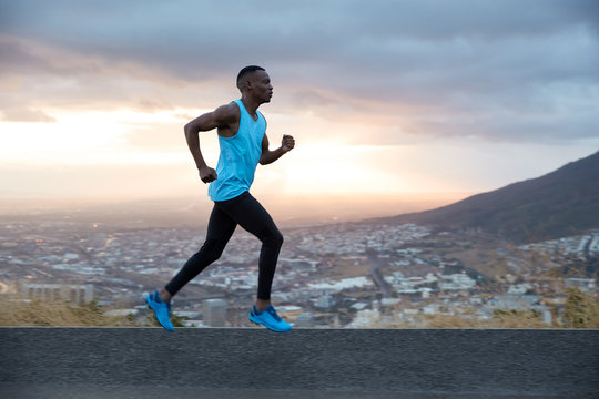 Full length horizontal shot of fast male runner enjoys speed, photographed in motion, runs along road with mountain and city view in background. Running at dawn concept. Strong sportsman outside