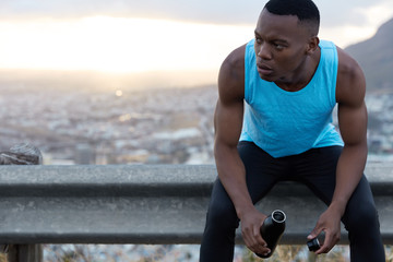 Male jogger rests after workout, holds bottle with refreshing drink, feels tired, poses over nature background with copy space area for your text message or information. Motivation, sport, endurance