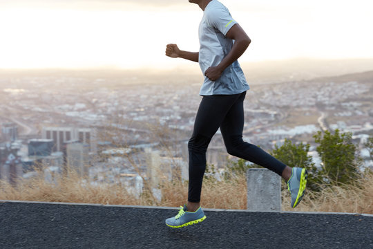 Cropped image of unrecognizable strong young man has morning jog across road, works on muscles, wears comfortable trainers, big city view is lower. People, sport, ethnicity and motion concept