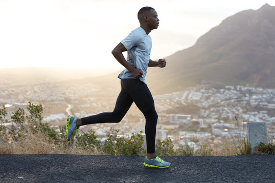 Outdoor shot of active dark skinned man joggs at morning, has regular trainings, dressed in tracksuit and comfortable sneakers, concentrated into distance, sees finish far away. Rock in background