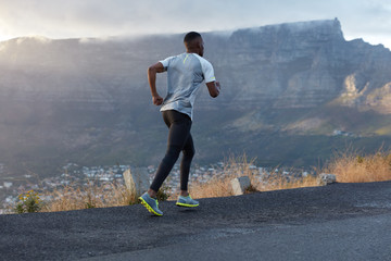 Fototapeta na wymiar Back shot of active dark skinned man being in action, runs across mountain road, leads healthy lifestyle, has endurance and motivation to be fit, poses over mountain background, enjoys nature