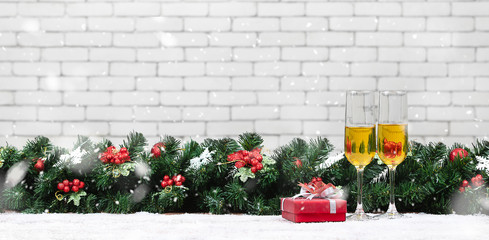 Fototapeta na wymiar Two champagne glasses setting next to a red gift box on snowy table decorate with pine branches, red holly berries. Copy space top area, blurred white brick wall background
