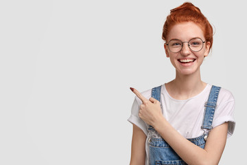 Studio shot of happy freckled lady in glasses, feels satisfied, wears dungarees, indicates at upper...
