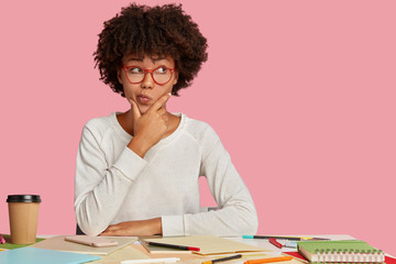 Doubtful student studies art, spends time on making sketches, holds chin and purses lips with...