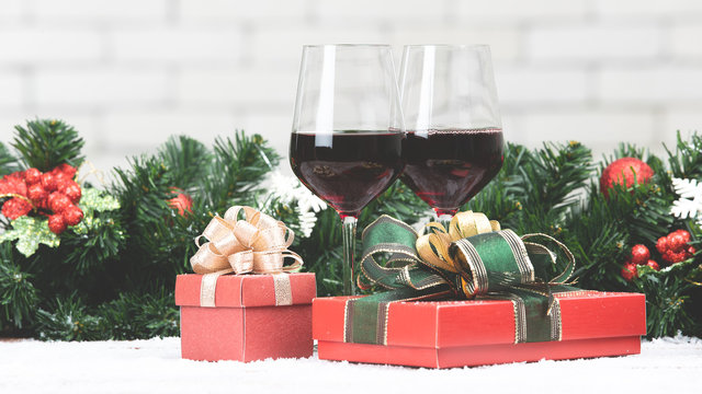 Two Red Wine Glasses Set Next To Red Gift Boxex On Snowy Table Prepare For Celabraion, Decorate With Pine Branches And Holly Berries. Blurred White Brick Wll Copy Space At Top Part