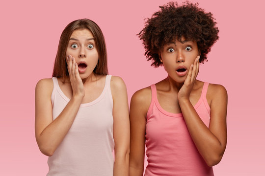 Waist up shot of puzzled frightened stupefied two girl of different nationalities, keep jaws dropped from surprisement, cant believe in astonishing news, model against pink background. Omg concept