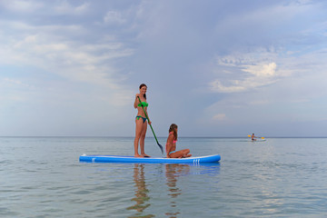Happy mother rolls her daughter on a Board for SAP surfing on the calm sea against the picturesque sky.