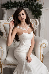 Fototapeta premium Portrait of a beautiful bride sitting on the white armchair. Hand in hair. Sharp cheekbones. Lace dress. Interior Studio.