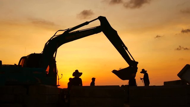 Construction Video At construction site The silhouette excavator is digging ground and lifting a large mortar, with engineering and construction workers to help with colorful sunset .