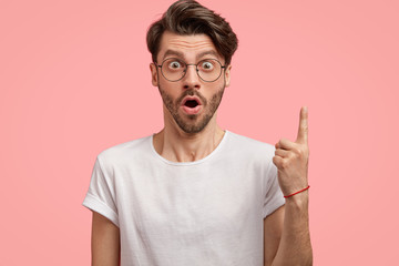 Isolated shot of surprised unshaven man gasps from amazement, points with fore finger upwards, wears round glasses and white t shirt, models against pink studio wall, demonstrates something above