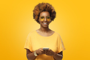Portrait of curly african american woman wearing disco style yellow glasses and t-shirt, holding smartphone, looking at camera and smiling