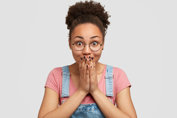Emotive pleased dark skinned female in eyewear covers mouth with both hands, sees something incredible, has crisp combed hair, models against white background. People, emotions, reaction concept