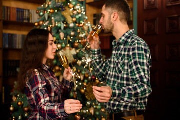 Merry Christmas and Happy New Year! Attractive young couple is celebrating holiday at home together, drinking champagne and smiling with Bengal lights in hand