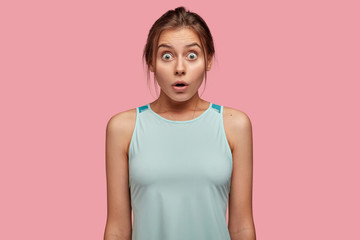 Photo of surprised girlfriend with bugged eyes full of fear, opens mouth widely, dressed in casual outfit, stands against pink background, expresses fear. Scared Caucasian sportswoman afraids
