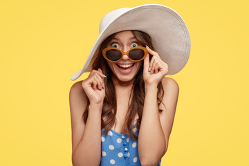 Fototapeta premium Horizontal shot of pretty attractive woman with dark hair, looks with surprise and happiness, keeps hand on rim of shades, models in summer outfit over yellow background. Beauty and emotions concept