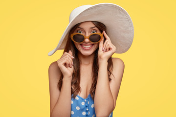 Image of good looking woman with satisfied expression, bright charming smile, looks through sunglasses, wears white hat, spends summer time in wonderful company of friends. Lady poses indoor