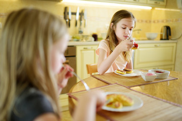 Cute little sisters enjoying their breakfast at home. Pretty children eating pancakes with strawberry sauce before school.
