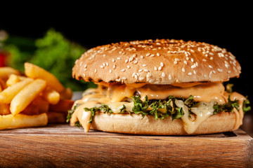 The concept of American cuisine. Juicy burger with meat patty, with cheese and french fries lying on a wooden board in a restaurant. Near a glass of beer with foam.