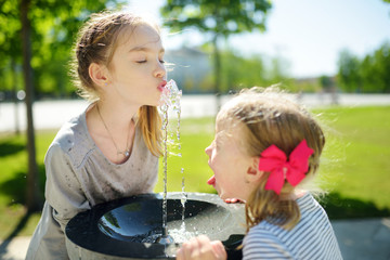 Two sisters having fun with drinking water fountain on warm and sunny summer day.