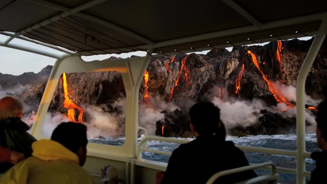 Tourists to Kilauea Hawaii volcanic area watching from boat as red hot lava falls into ocean causing rising steam seen from distance 