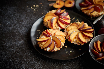 Delicious homemade mini tarts with fresh sliced plum fruit. Sprinkled with icing sugar. Placed on black ceramic plates with fresh pieces of plums on sides. Dark rusty background from top.