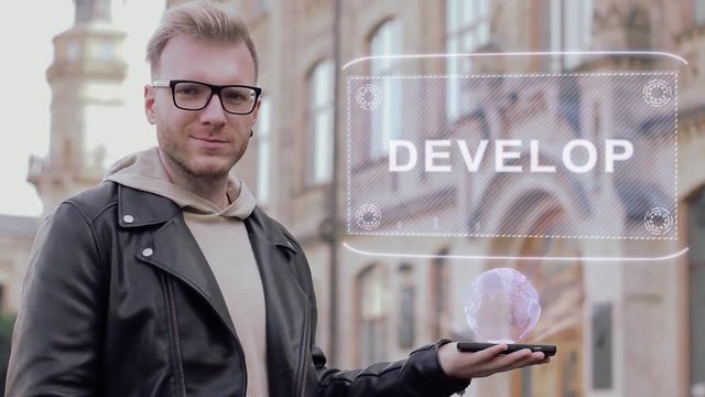 Smart young man with glasses shows a conceptual hologram Develop. Student in casual clothes with future technology mobile screen on university background