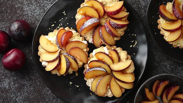 Delicious homemade mini tarts with fresh sliced plum fruit. Sprinkled with icing sugar. Placed on black ceramic plates with fresh pieces of plums on sides. Dark rusty background from top.