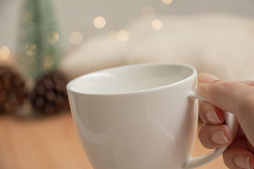 white cup of hot chocolate on table with blur christmas light,pine cones,mini tree and sweater.