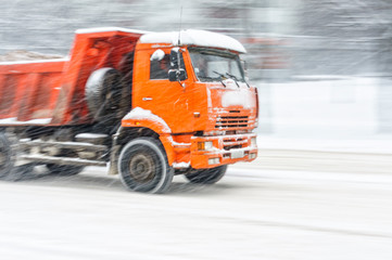 Big orange dump truck rides on a snowy road during a snow storm. Blurred in motion background.