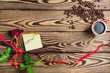 Inscription call me on paper beside red rose with ribbon and cup of coffee near coffee beans on old wooden table. Top view with copy space. Saint Valentine's Day concept