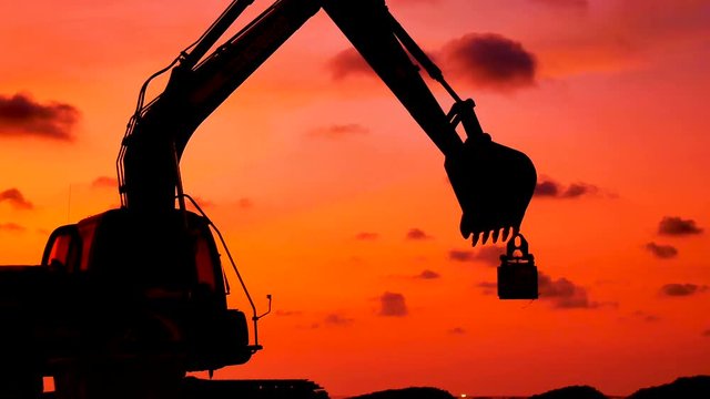 Construction Video At construction site close up The silhouette excavator is digging ground and lifting a large mortar, with engineering and construction workers to help with colorful sunset .