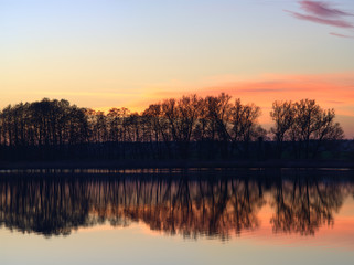 Orange and pink sunset rural landscape with row of trees, reflecting in the tranquil waters of a lake in the german countryside.