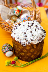 Easter cake with white and black chocolate sauce, spring flowers and an egg on a bright yellow tablecloth, behind an Easter basket